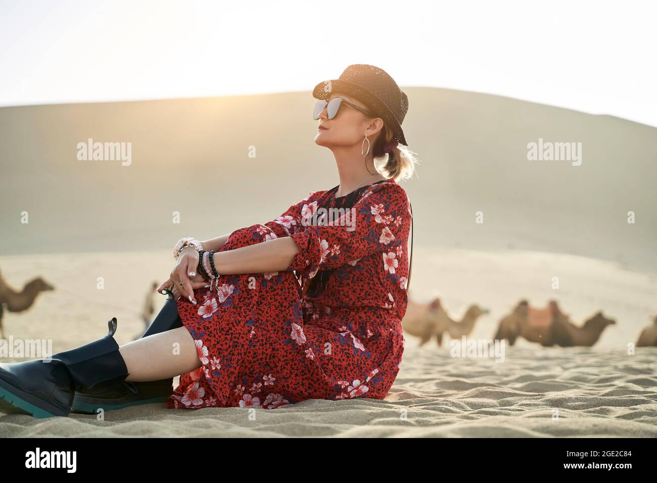 donna asiatica in abito rosso seduta nel deserto guardando la vista con caravan di cammelli e enorme duna di sabbia sullo sfondo Foto Stock