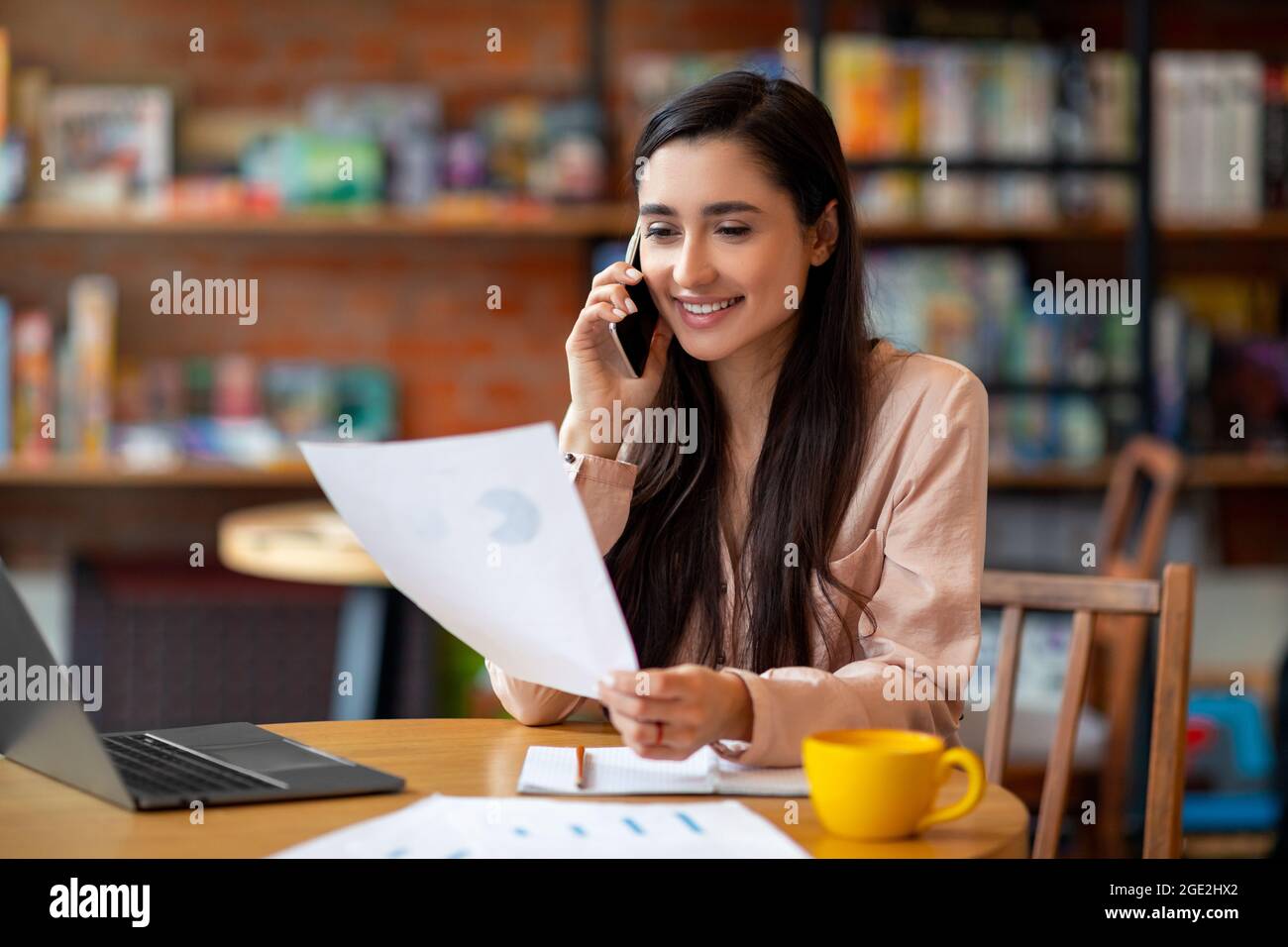 Ufficio remoto. Felice donna latina che lavora con documenti e parla con il cliente sul cellulare, seduto all'interno del bar Foto Stock