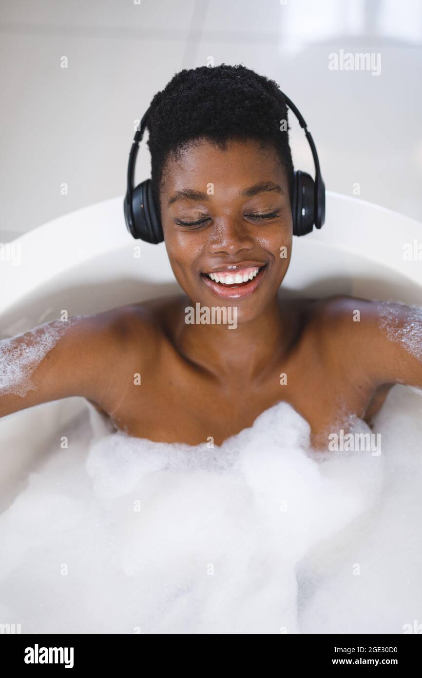 Donna afroamericana sorridente che si rilassa in bagno con gli occhi chiusi ascoltando musica su cuffie Foto Stock