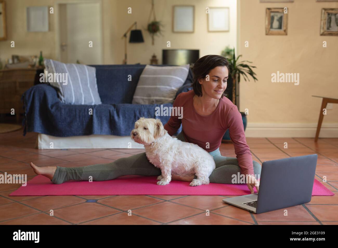 Sorridendo donna caucasica in soggiorno con il suo cane da compagnia, praticando yoga, usando il computer portatile Foto Stock