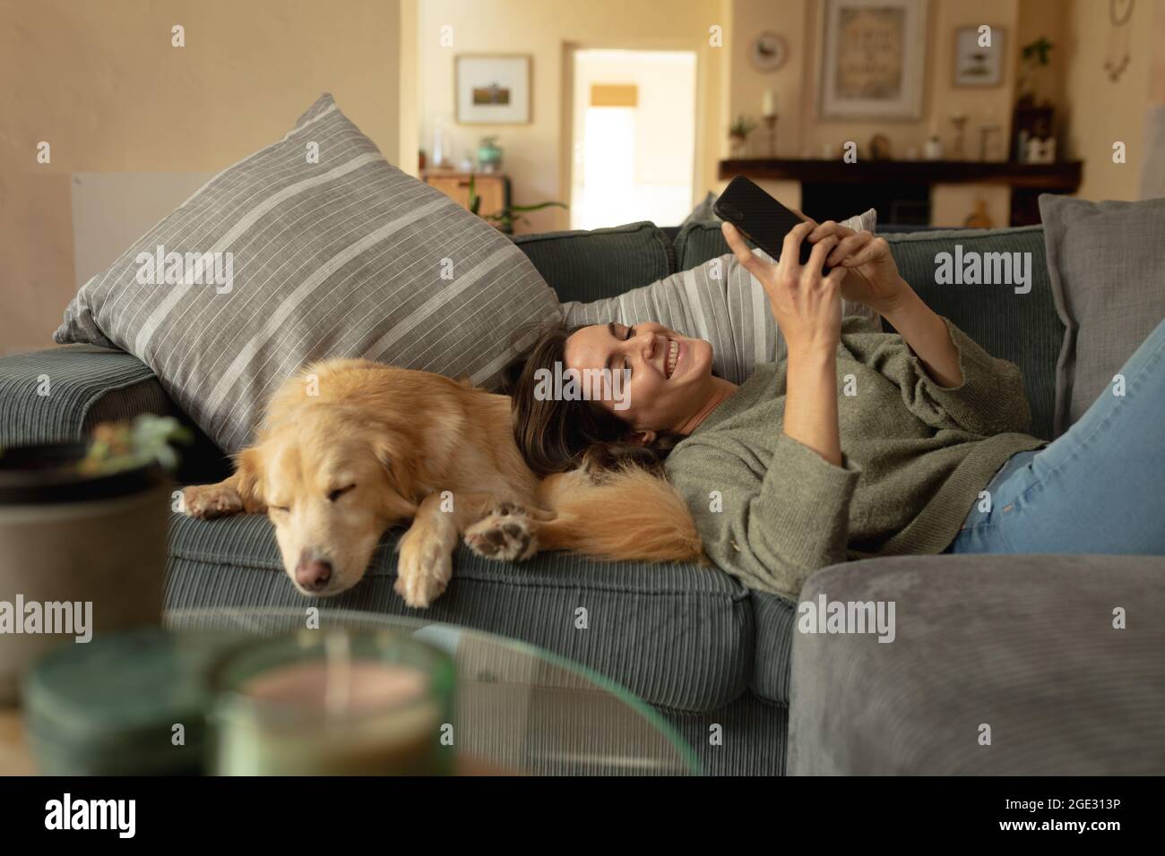 Sorridente donna caucasica in soggiorno, sdraiata sul divano con il suo cane da compagnia, usando lo smartphone Foto Stock