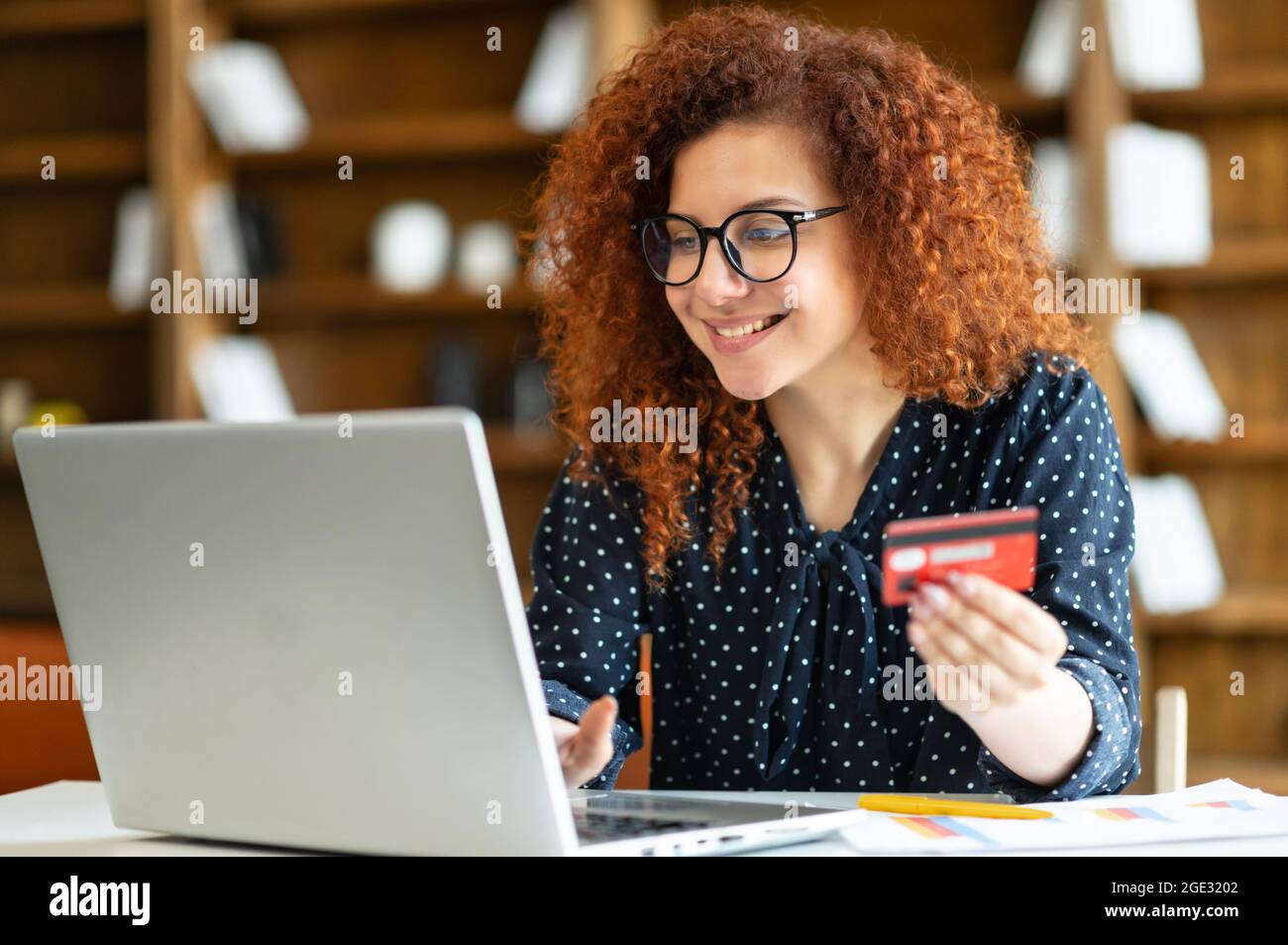 Donna d'affari sorridente con acconciatura curly che tiene la carta di credito per fare la transazione in linea, guardando il laptop, l'acquisto degli articoli desiderati, facendo l'acquisto in linea, shopping dal concetto domestico Foto Stock