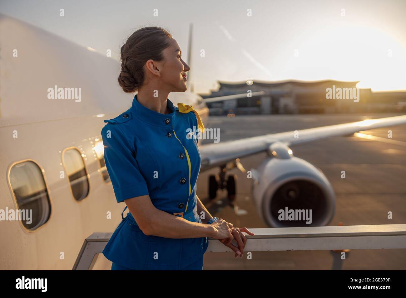 Aria premurosa in uniforme blu che guarda lontano, in piedi all'aperto al tramonto. Aereo commerciale vicino al terminal in un aeroporto nel Foto Stock
