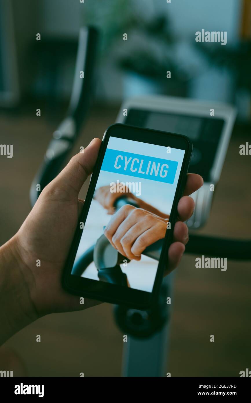 primo piano di un giovane caucasico che guarda al suo smartphone, che legge il ciclismo sul suo schermo, di fronte ad una bicicletta stazionaria nel soggiorno di h. Foto Stock