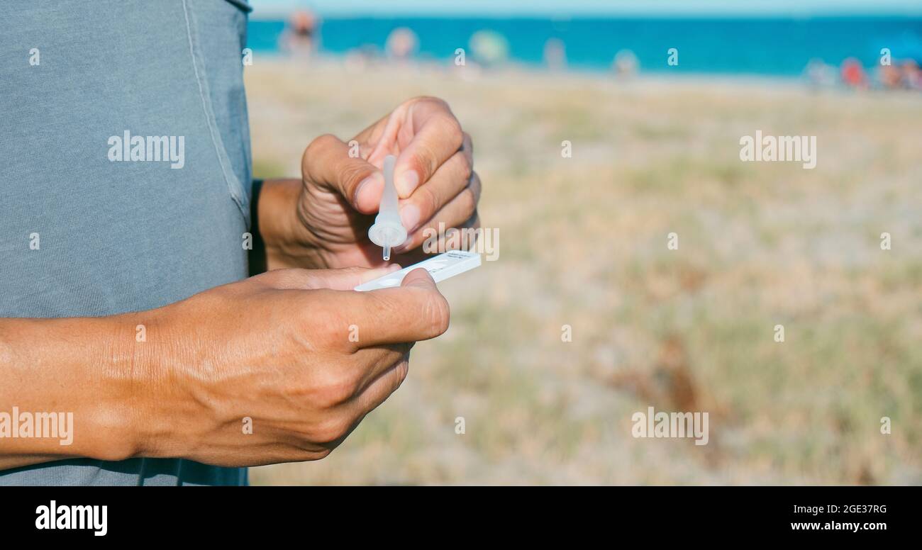 primo piano di un giovane caucasico sulla spiaggia, in abbigliamento casual, mettendo il proprio campione nel covid-19 antigen diagnostic test device, in una panoramica Foto Stock