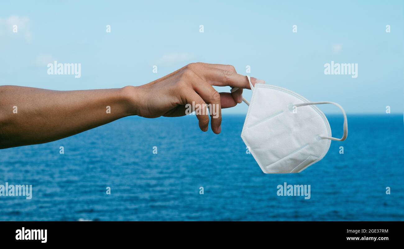Un giovane caucasico tiene in mano una maschera FFP2 bianca di fronte all'oceano, mentre lo indossa in un formato panoramico Foto Stock