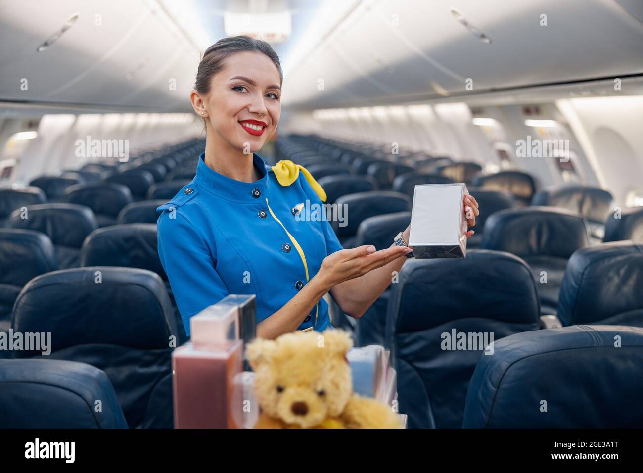 Abbastanza stewardess in uniforme blu brillante sorridente alla macchina fotografica, che fa pubblicità al profumo come regalo mentre conduce il carrello del carrello attraverso la corsia dell'aereo vuota Foto Stock