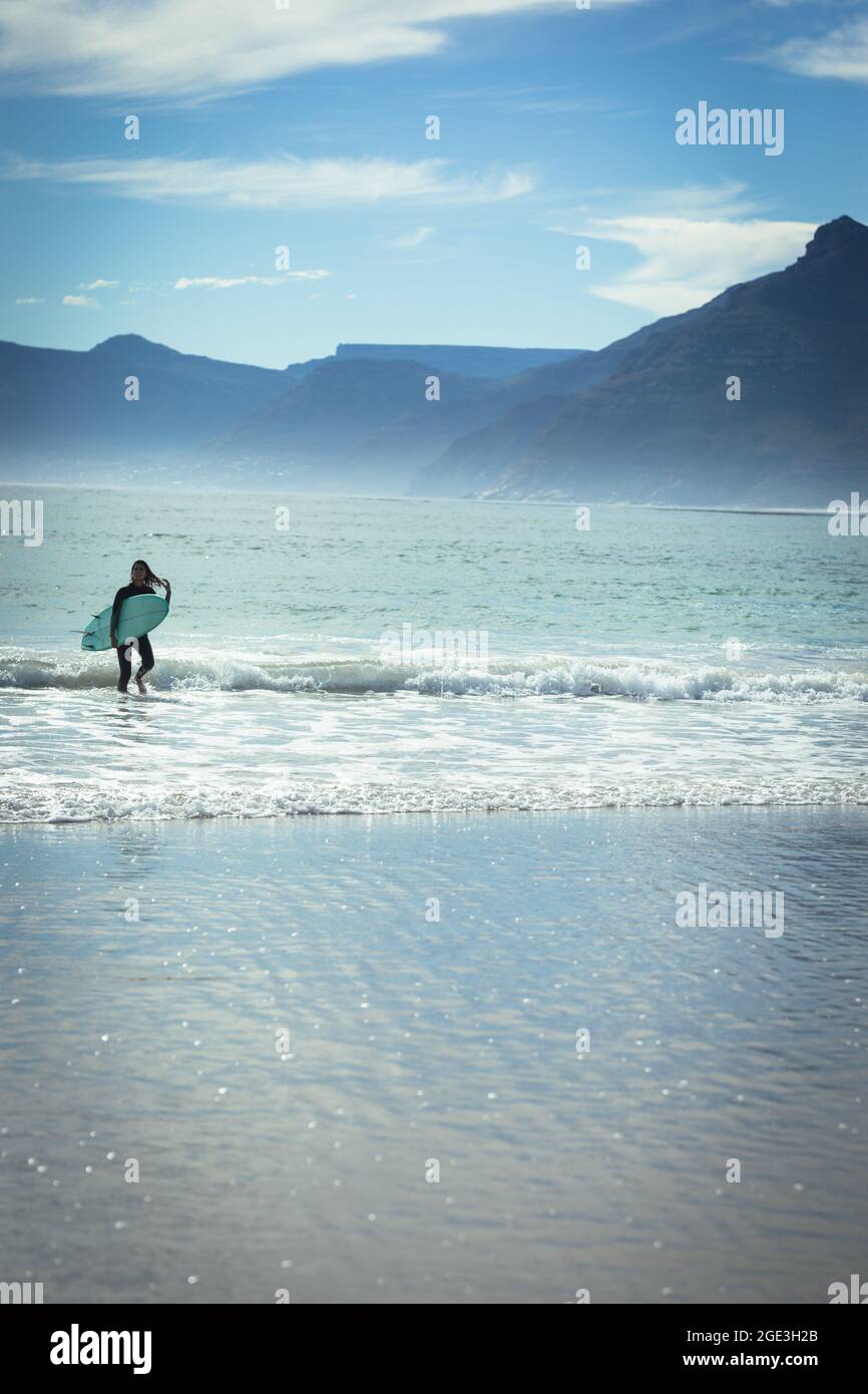 Razza mista donna a piedi e tenendo surf in mare in giorno di sole Foto Stock