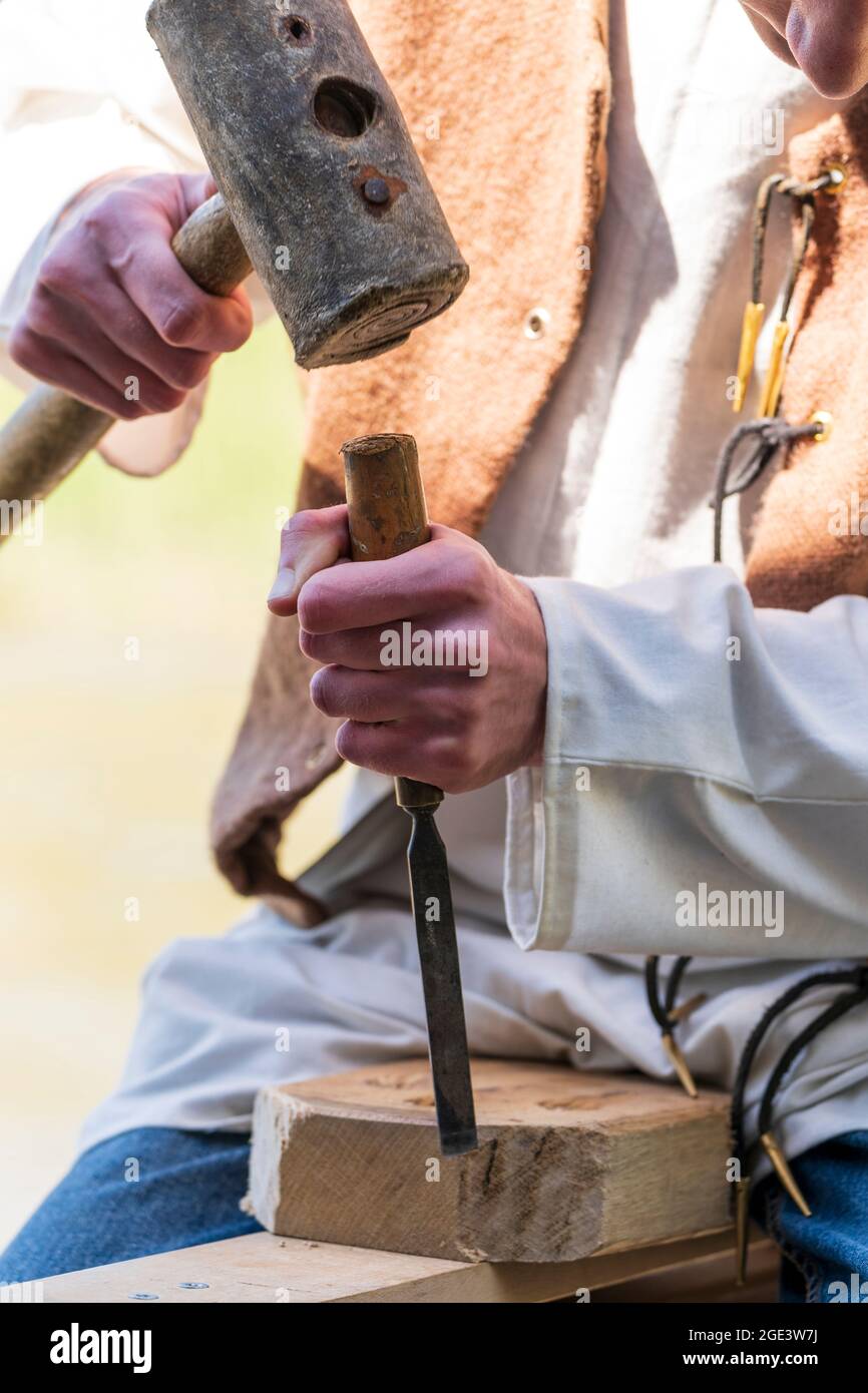 Falegname medievale maschile reenactor. Primo piano di un uomo che tiene un mazzuolo di legno che colpisce uno scalpello contro un blocco di legno. Foto Stock