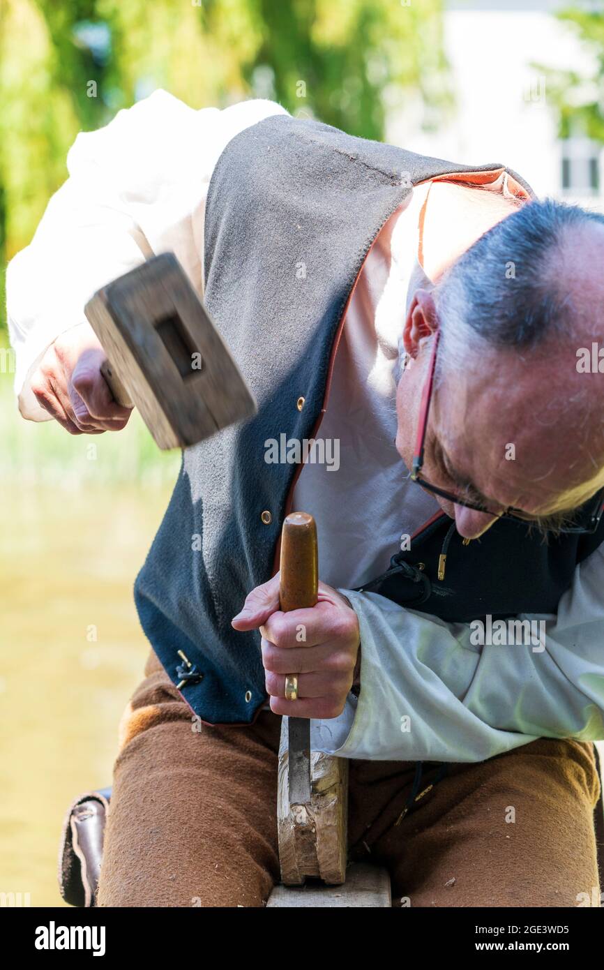 Primo piano di un uomo anziano, carpentiere medievale vivente storia reenactor, utilizzando mazzuolo e scalpello per modellare un po 'di legno con sfondo verde fogliame. Foto Stock