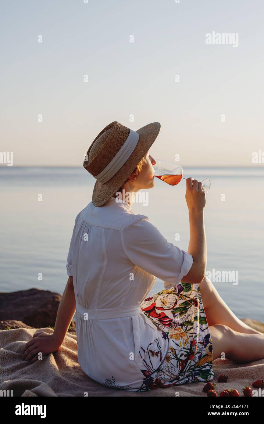 Giovane donna in cappello di paglia che beve vino sulla riva del mare al  tramonto Foto stock - Alamy