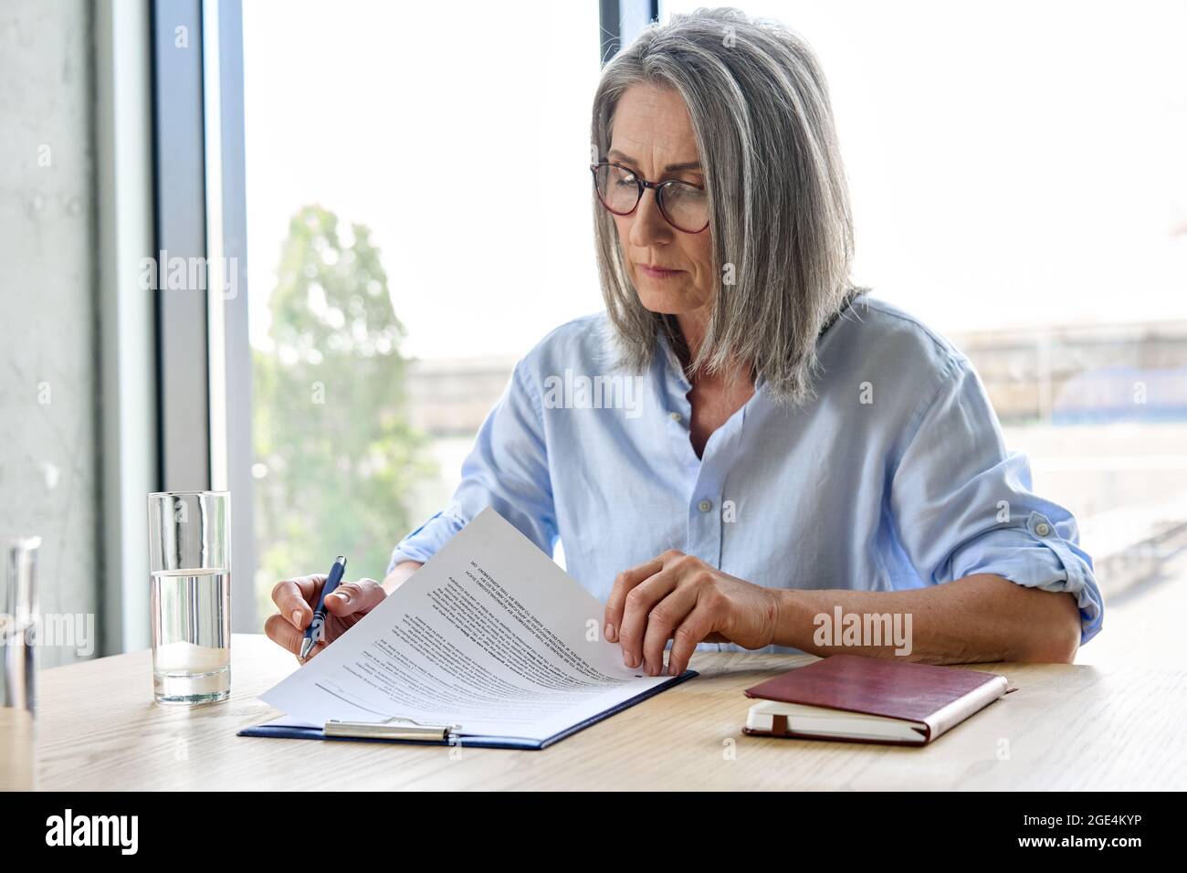 Donna d'affari anziana seria in occhiali che legge il contratto di partenariato a tavola. Foto Stock