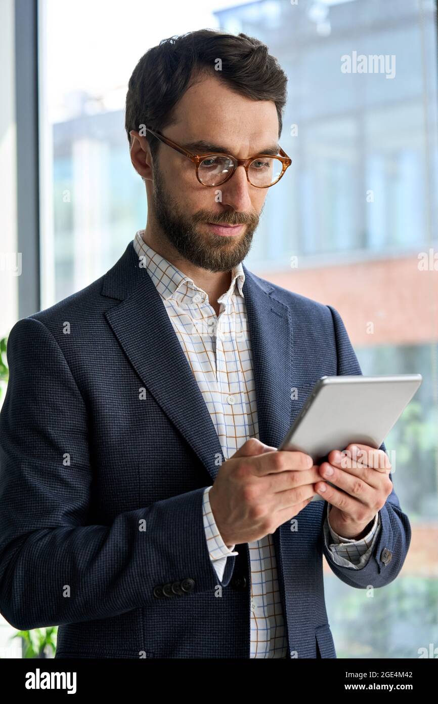 Uomo d'affari serio e maturo che utilizza un dispositivo tablet in piedi in ufficio. Foto Stock
