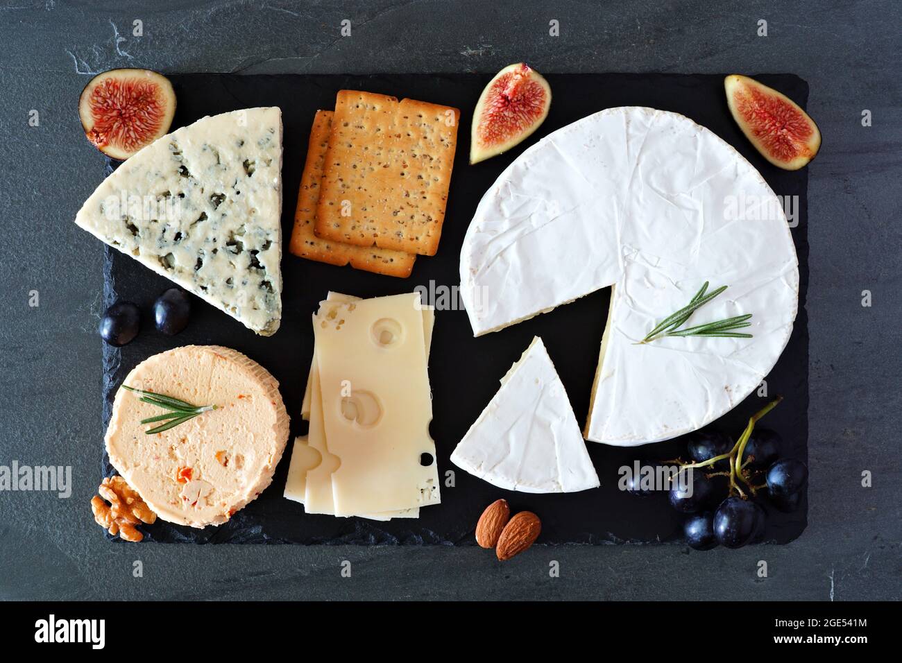 Tavola di formaggio con una selezione di formaggi, cracker, fichi e noci su tavola di servizio di ardesia. Sopra la vista su uno sfondo scuro. Foto Stock