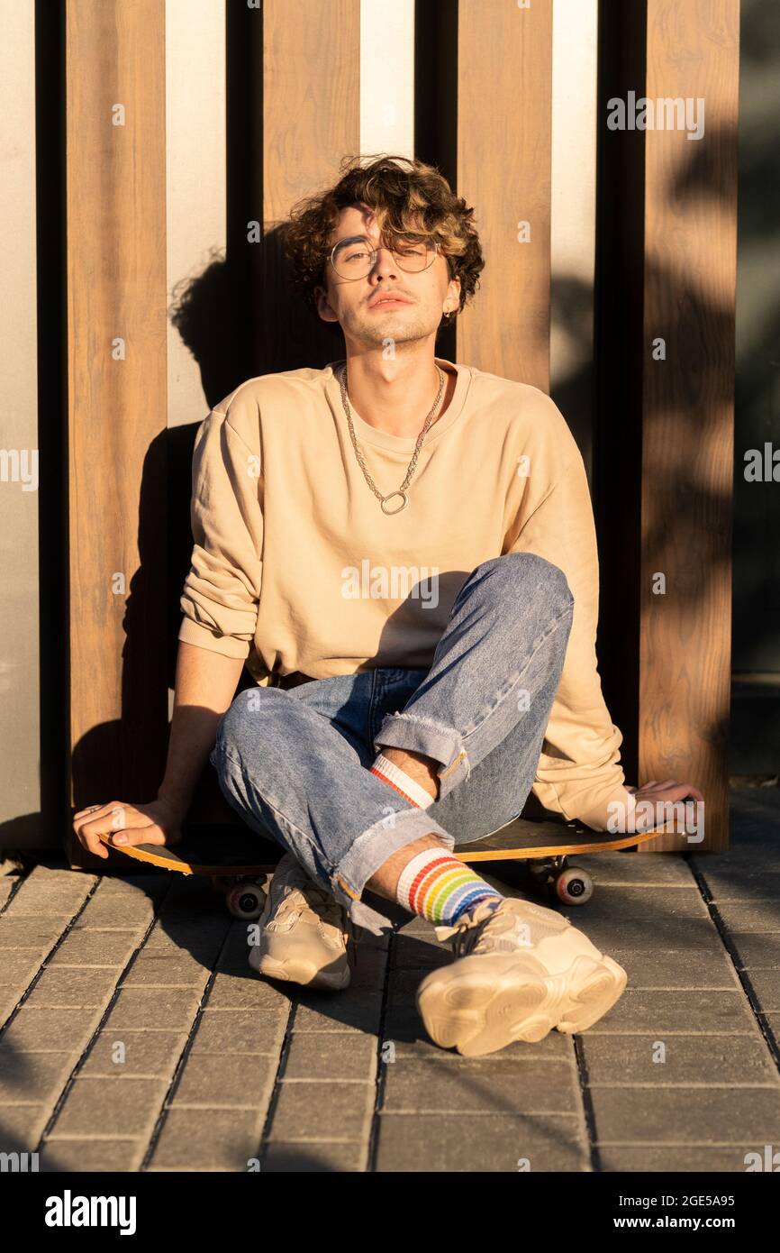 Ragazzo rilassato in casualwear seduto a parete sul marciapiede e godendo di una giornata di sole Foto Stock