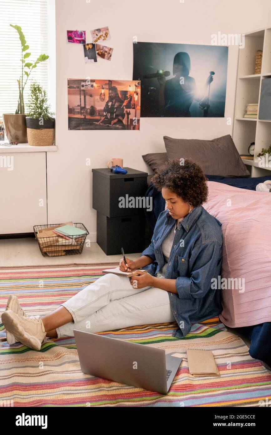Adolescente ragazza in casualwear che effettua l'assegnazione domestica mentre si siede sul pavimento della camera da letto Foto Stock