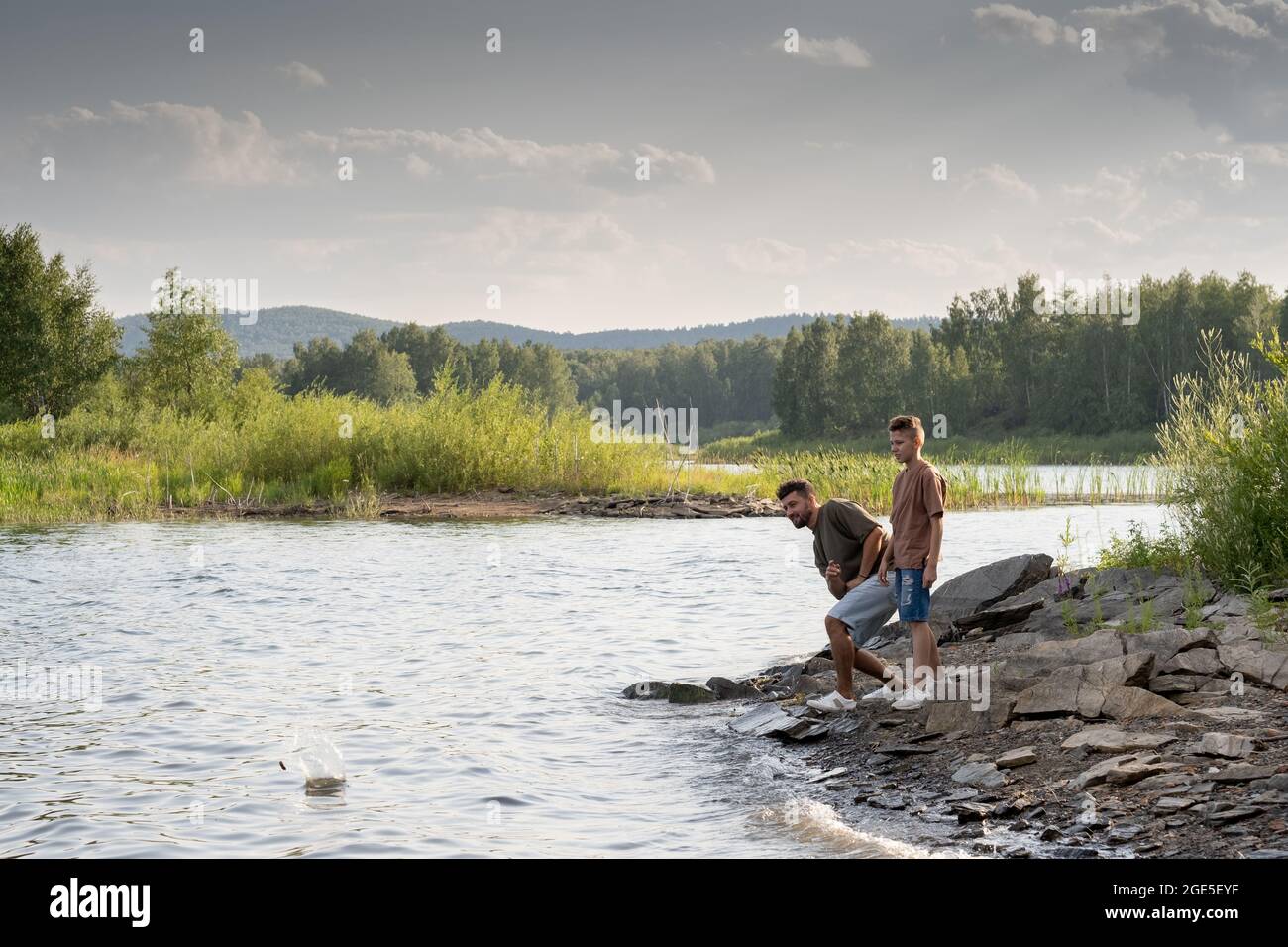 Giovane uomo e ragazzo adolescente godendosi il tempo vicino al lago e gettando piccole pietre in acqua Foto Stock
