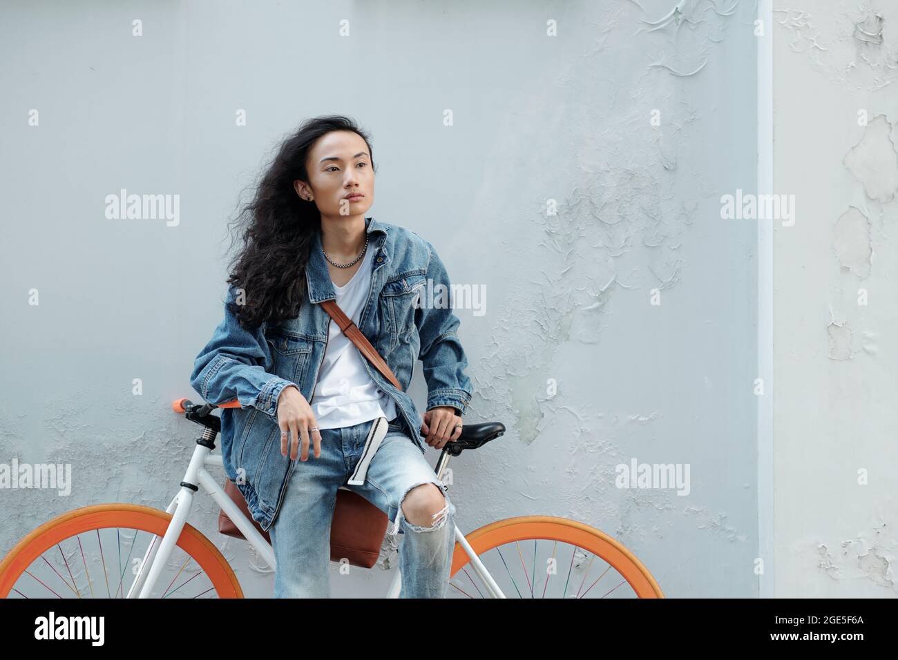 Elegante giovane in jeans e giacca in denim che pende in bicicletta e guarda lontano Foto Stock