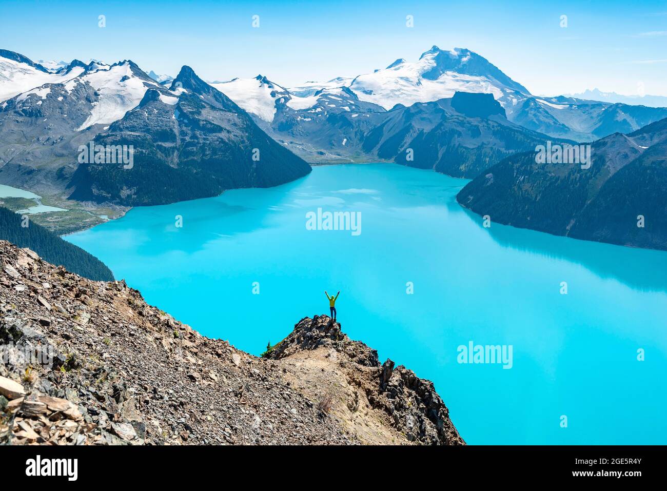 Giovane uomo in piedi su una roccia, stendendo le braccia nell'aria, vista delle montagne e del ghiacciaio con il lago blu turchese Garibaldi Lago, cime Panorama Ri Foto Stock