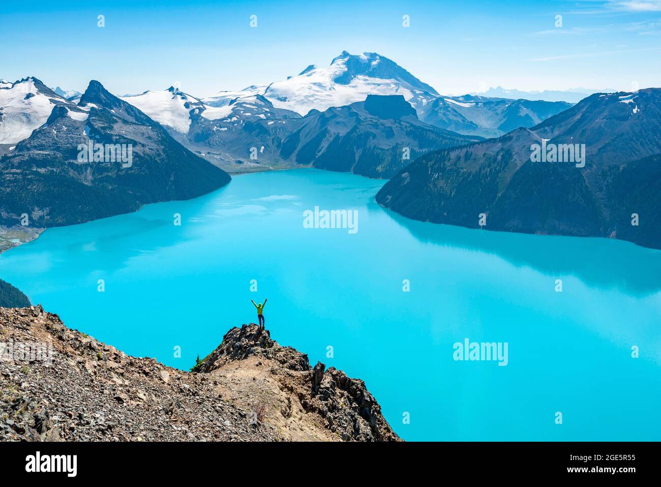 Giovane uomo in piedi su una roccia, stendendo le braccia nell'aria, vista sulle montagne e ghiacciaio con il lago blu turchese Garibaldi Lago, Peaks Panorama Foto Stock