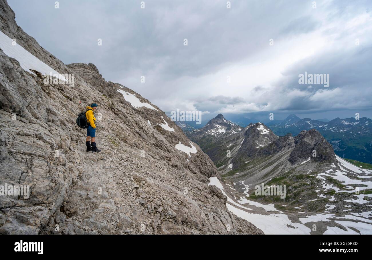 Escursionista su sentiero escursionistico in terreno roccioso, panorama montano con vecchi campi di neve e cime rocciose di montagna sullo sfondo, drammatico cielo nuvoloso Foto Stock