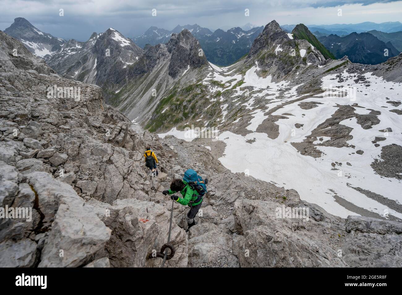 Escursionista e escursionista discesa terreno roccioso, percorso fissato con corda d'acciaio, sullo sfondo panorama montano con vecchi campi di neve e montagna rocciosa Foto Stock