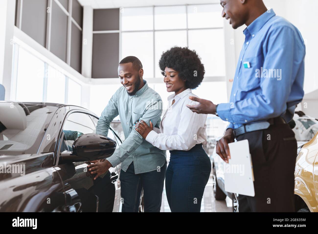 Vendite di automobili. Il manager parla con la coppia afro, mostrando loro la nuova auto presso il negozio della concessionaria. Giovane famiglia nera che sceglie il veicolo, guardando moderno Foto Stock