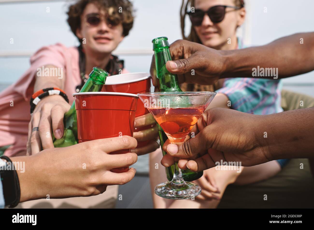 Mani di amici che tostano con bottiglie di birra, tazze di plastica e bicchieri martini alla festa Foto Stock