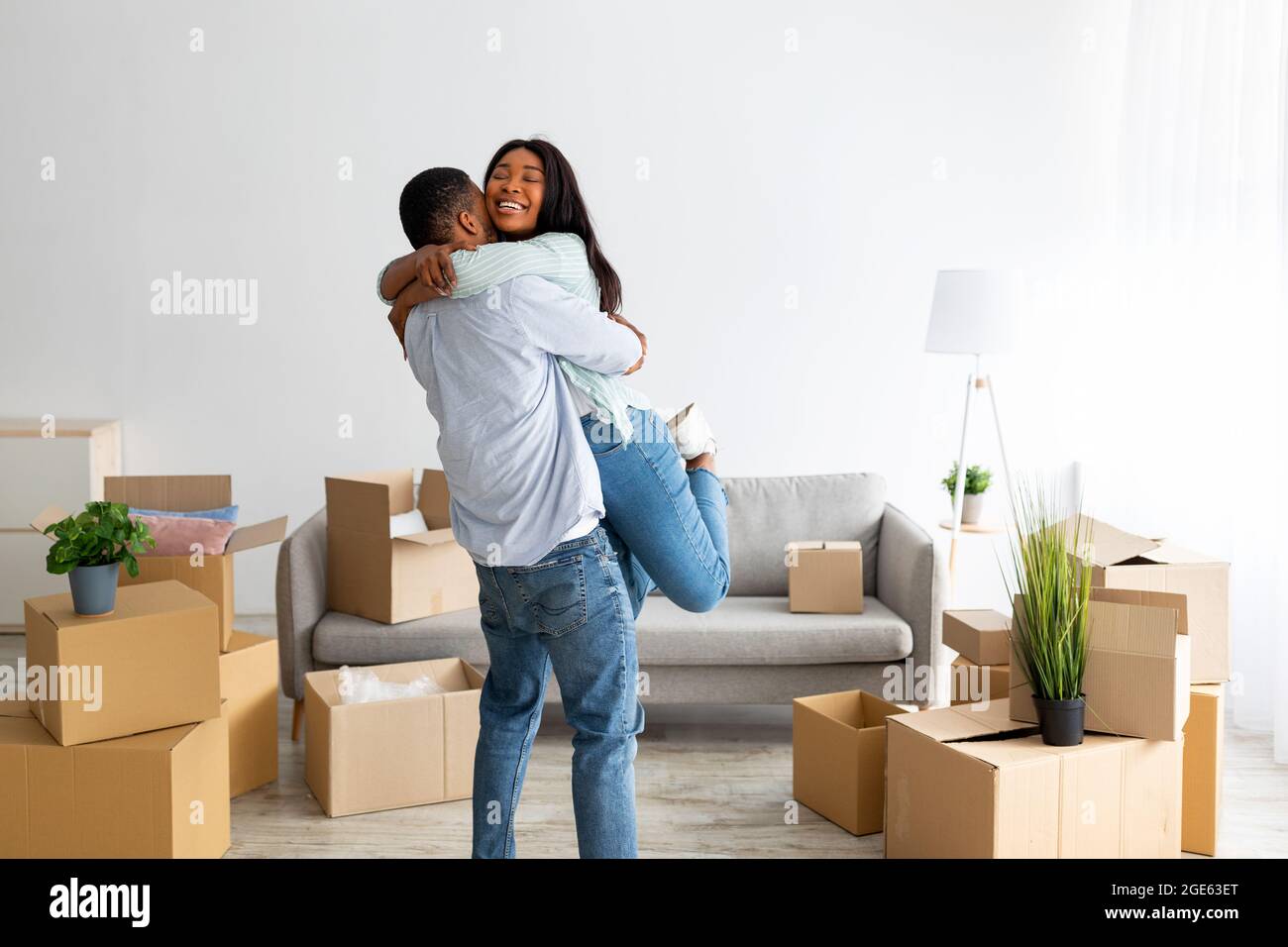 Proprietari di casa. Felice uomo afroamericano abbracciare e sollevare moglie, festeggiando giorno in movimento nel loro nuovo appartamento, spazio libero. Giovani inquilini neri feeli Foto Stock