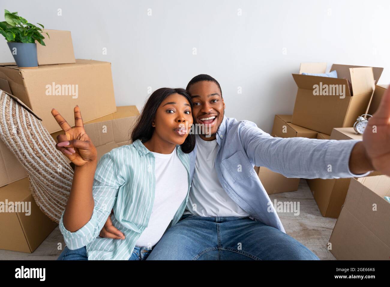 Felici proprietari di casa. Giovane coppia afro-americana che prende selfie e gesturing segno di vittoria dopo essersi trasferito in nuova casa e seduto tra scatole di cartone, Foto Stock