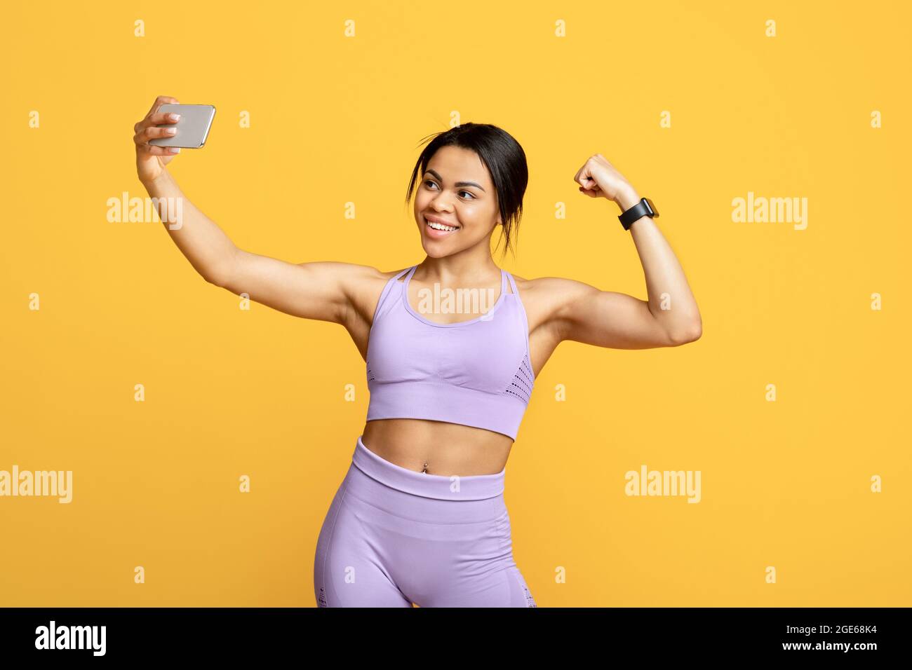 Selfie dal blogger del formatore. Felice fit donna nera in sportswear facendo foto su smartphone per blog e mostrando il suo bicipite, in posa sul backgrou giallo Foto Stock