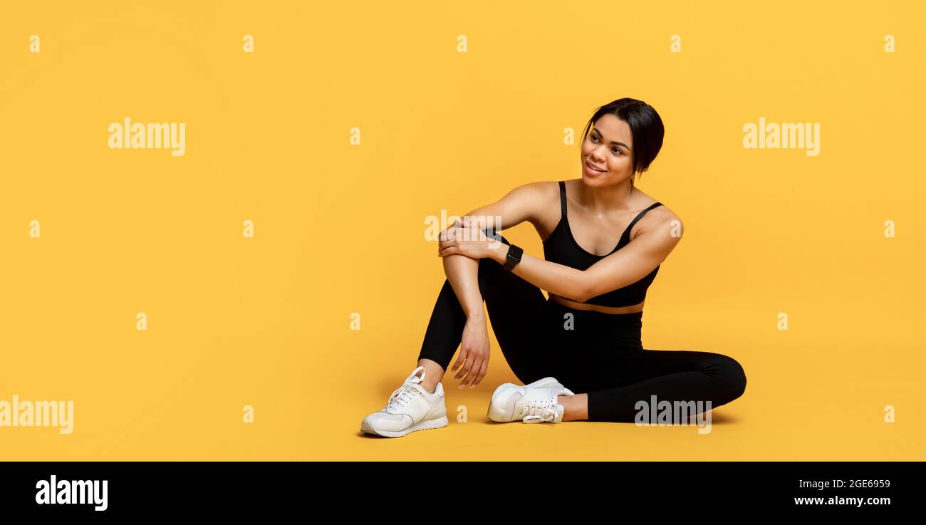 Atletica donna afroamericana che riposa dopo l'allenamento, seduto sul pavimento a sfondo giallo studio e guardando da parte lo spazio vuoto. Fitness e healt Foto Stock
