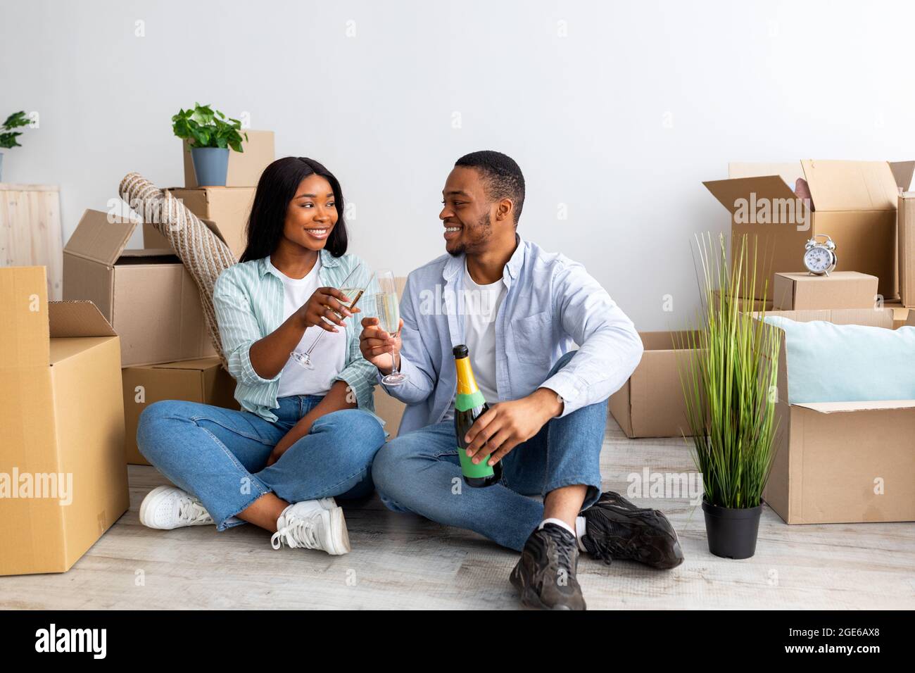 Giovane coppia afroamericana festeggiando il trasferimento al nuovo appartamento, seduto tra scatole di cartone e bere champagne. Proprietari di casa e case di famiglia Foto Stock