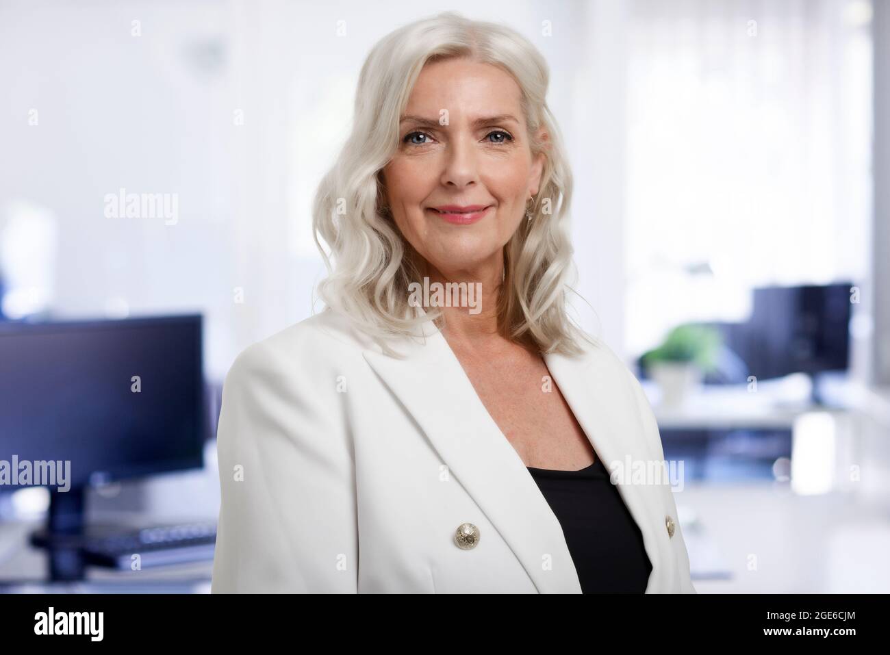 Ritratto di bella donna d'affari anziana con capelli grigi in piedi in ufficio mentre si guarda la macchina fotografica e sorridente. Foto Stock