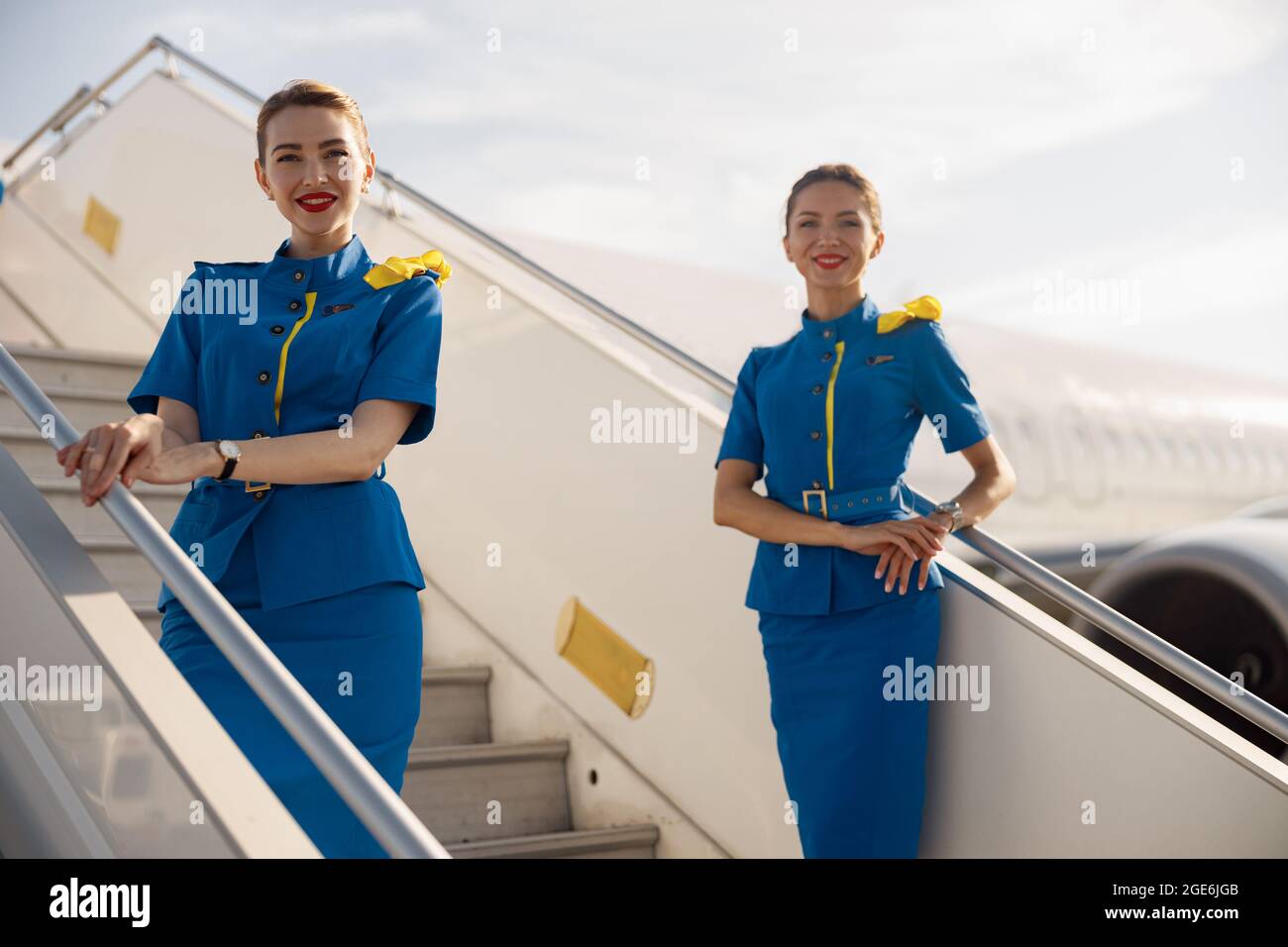 Due bellissime camere d'aria in uniforme blu che sorridono alla telecamera, in piedi su un'aerostazione e accogliendo i passeggeri Foto Stock