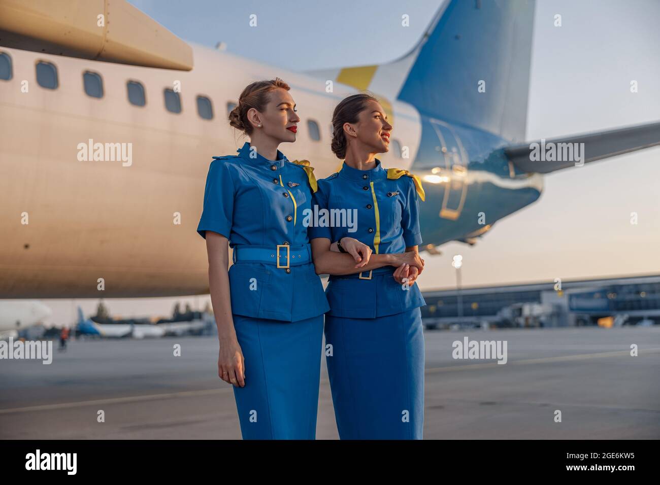 Due belle hostess in uniforme blu sorridenti, in piedi di fronte a un grande aereo passeggeri in aeroporto al tramonto Foto Stock