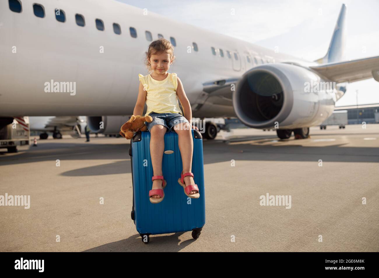 Foto a lunghezza intera di carina bambina che tiene il suo giocattolo, guardando la macchina fotografica e seduto sulla valigia di fronte al grande aereo Foto Stock