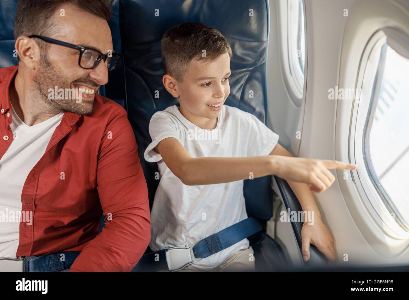 Felice ragazzino che guarda fuori la finestra, mostrando qualcosa al padre mentre si siede vicino alla finestra, viaggiando in aereo insieme Foto Stock