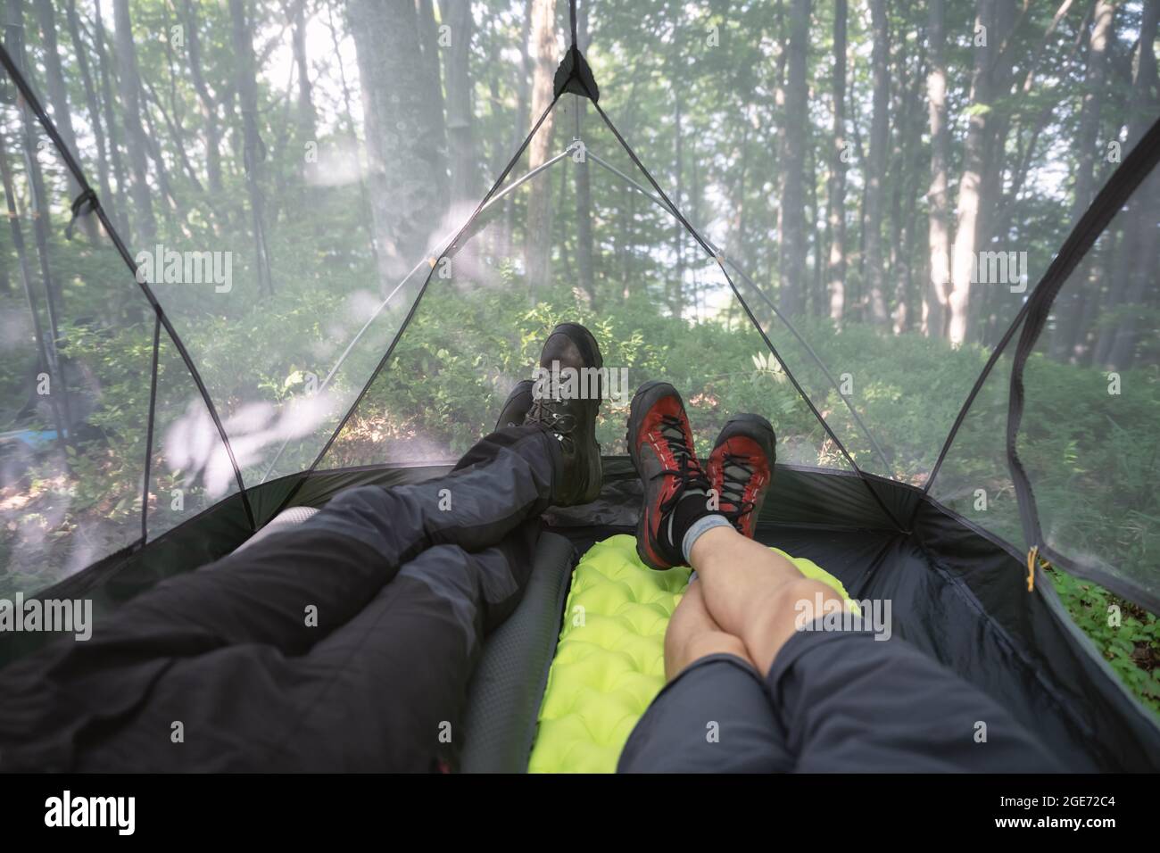 Le gambe maschili all'interno della tenda da campeggio con la foresta estiva sullo sfondo. Due viaggiatori che si trovano all'interno della tenda turistica e godono la vista della natura bella. Concetto di viaggio, escursioni e campeggio Foto Stock