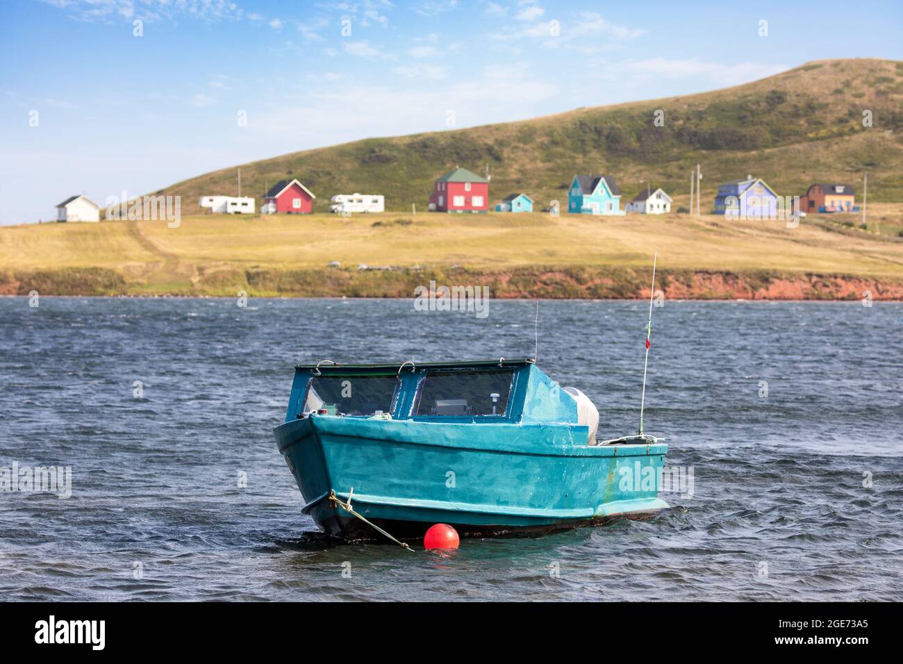 Piccola barca blu ormeggiata nelle acque blu delle isole Magdalene, Canada. Case colorate in legno fiancheggiano la costa dietro. Foto Stock