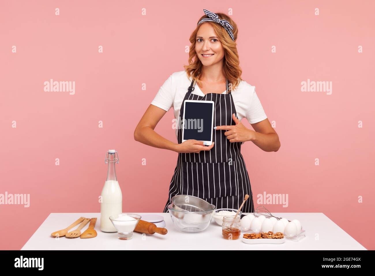 Ritratto di bella giovane donna adulta che tiene il computer tablet e che punta verso lo schermo vuoto mentre si sta in piedi vicino al tavolo con ingredienti da forno. IND Foto Stock