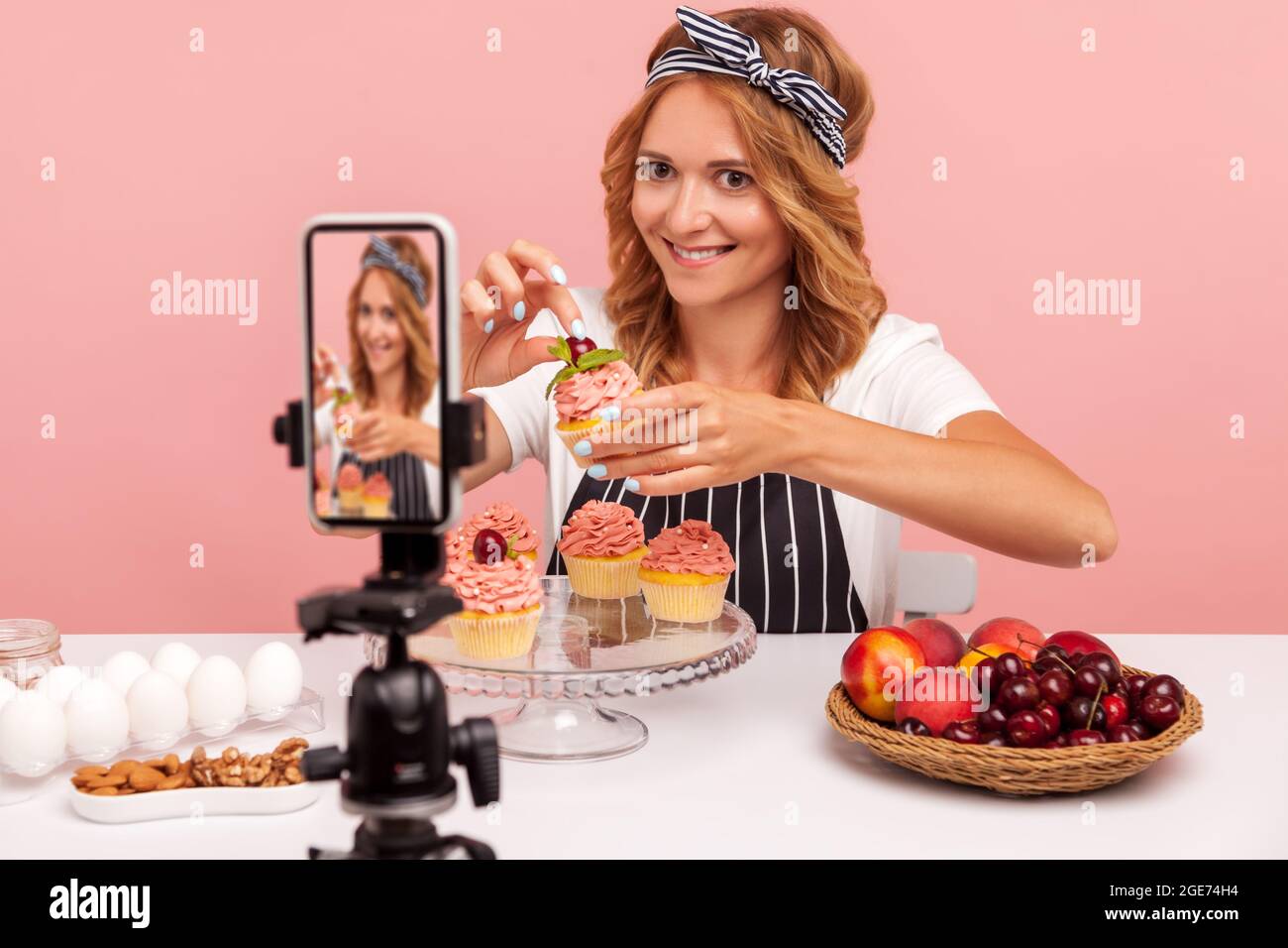 Food vlogger tenere piccola torta decorando con crema e ciliegia, guardando la fotocamera su smartphone su treppiede e sorridendo, registrare video per vlog. Poll Foto Stock