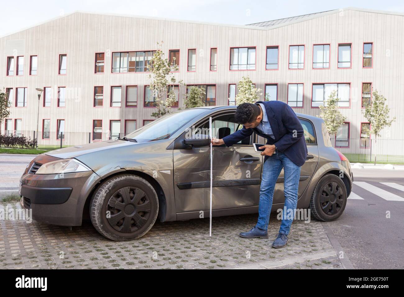 Vista laterale della bruna maschio auto regolatore di assicurazione ispezionando un veicolo che è stato in un relitto incidente, indossando jeans e giacca, valuta la diga Foto Stock