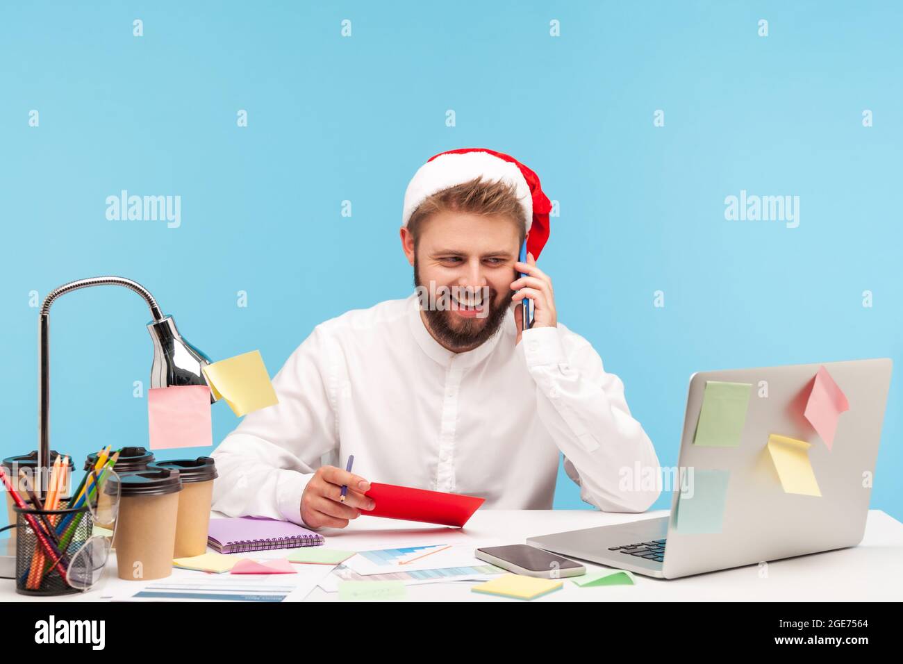 Uomo sorridente e positivo con barba in babbo natale smartphone parlante tenendo in mano la busta rossa con lettera e cartolina, seduto sul posto di lavoro. Poll Foto Stock