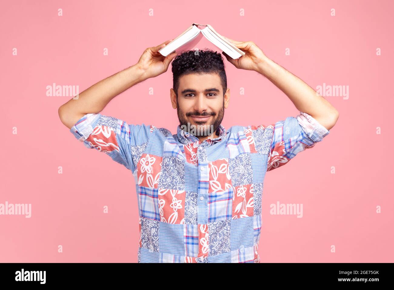 Ritratto di uomo con barba in camicia blu casual stile tenendo aperto il libro sulla testa, guardando la macchina fotografica con sorriso, esprimendo emozioni positive. Interno Foto Stock