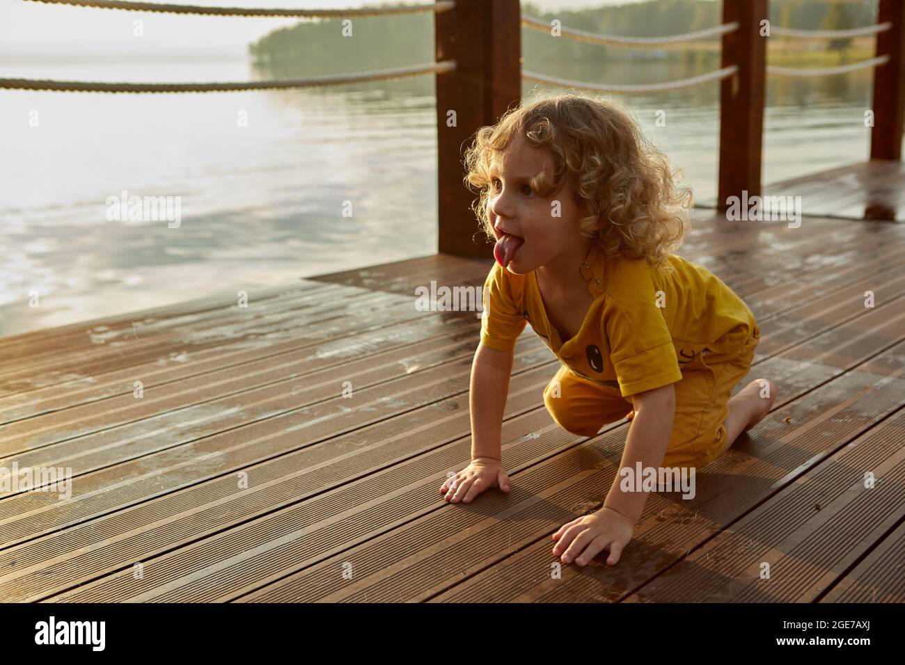 Adorabile toddler con capelli ricci che mostrano la lingua e strisciando sulla banchina bagnata vicino all'acqua del lago al tramonto Foto Stock