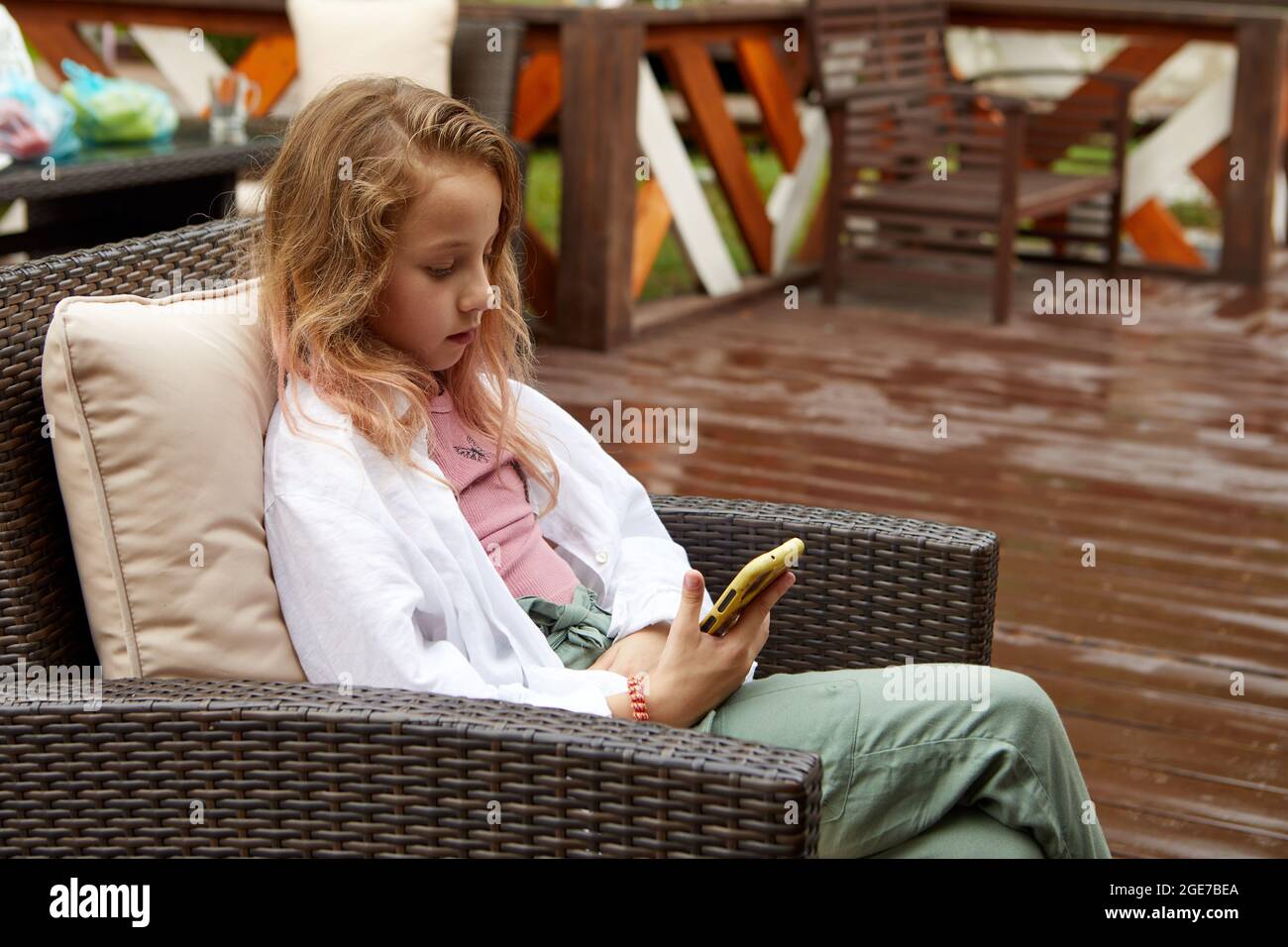 Presteen ragazza in vestiti casual appoggiata sul cuscino e guardando video sul cellulare mentre si siede sulla poltrona di vimini sulla terrazza Foto Stock