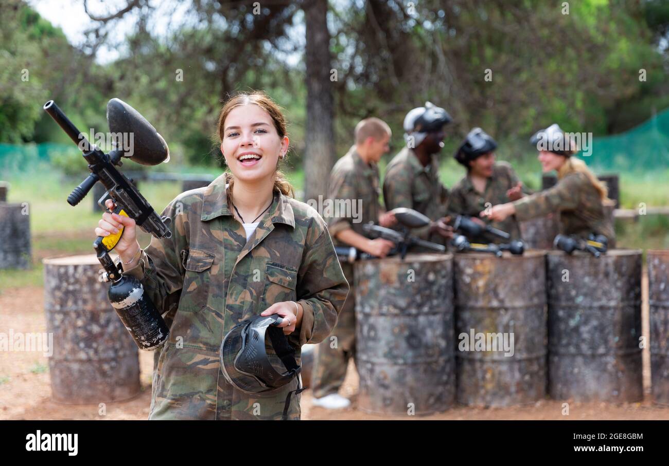Giovane giocatore di paintball femminile in camouflage in piedi sul campo di battaglia con la pistola prima di giocare Foto Stock