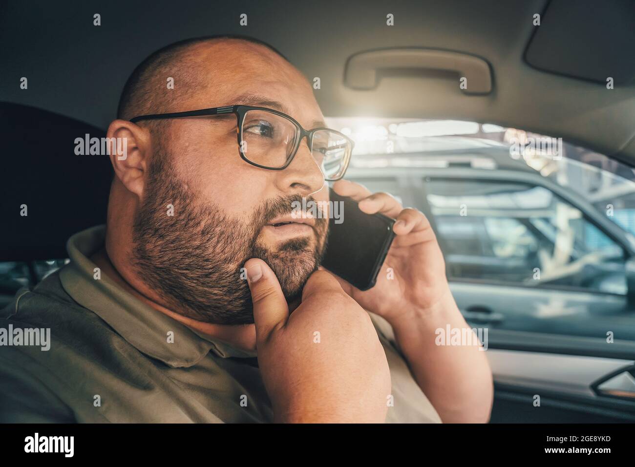 Uomo perplesso con occhiali che parlano al telefono mentre si guida in auto. Emozione di dubbio e diffidenza sul volto di divertente uomo grasso. Foto Stock