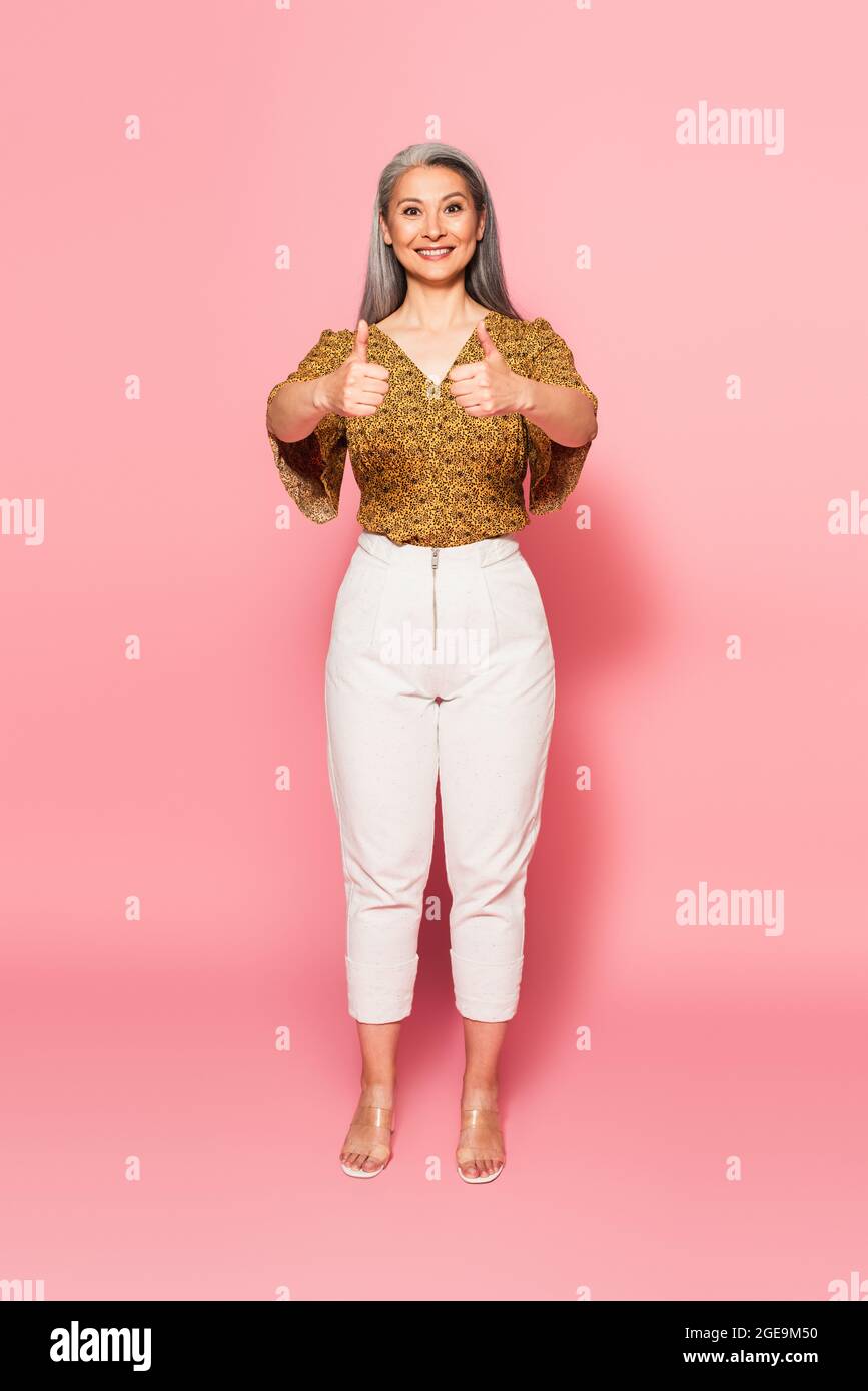 vista completa della donna matura in blusa a motivi geometrici e dei pantaloni  bianchi che mostrano i pollici sul rosa Foto stock - Alamy